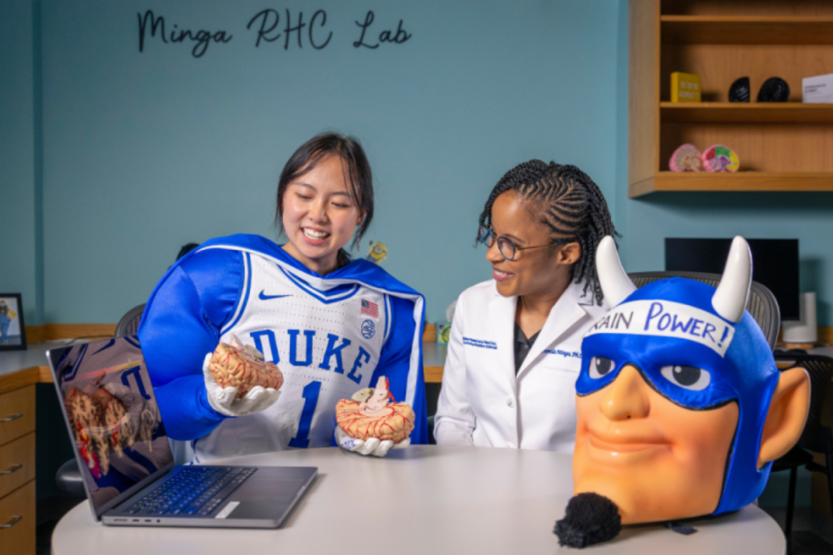 Nana osaki wears the duke blue devil mascot uniform without the head. She has a brain in her hand is sitting at a table with Dr. Jamila Minga. The blue devil mascot head says "brain power" and sits on the table.
