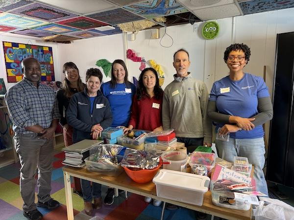 seven people standing behind a table filled with school supplies